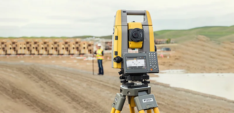 Total station presisi di atas tripod menghadap lokasi konstruksi, dengan surveyor bekerja di kejauhan